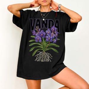 May include: Black t-shirt featuring the word "VANDA" in purple block letters above a detailed illustration of purple Vanda orchids. The graphic includes green leaves, roots, and flowers, creating a botanical design.