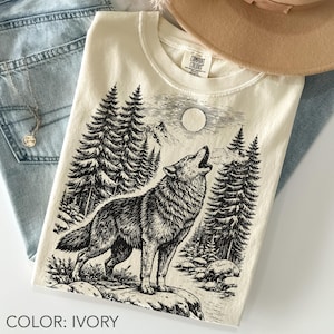 Ivory Comfort Colors unisex oversized t-shirt featuring a vintage black line art wolf howling beneath a full moon, surrounded by pine trees, rocky mountains, forest plants, and wilderness scenery.