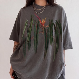May include: A charcoal gray short-sleeve t-shirt featuring a large, detailed botanical illustration of a philodendron plant with long, dark green leaves and a single vibrant orange and purple bird of paradise flower centered on the chest.