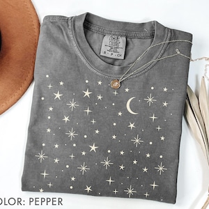 Puede incluir: Una camiseta gris Comfort Colors con un diseño celestial. La camiseta presenta un patrón de estrellas y una luna creciente en color crema. Un collar plateado con un colgante está drapeado sobre la camiseta. La camiseta está etiquetada "COLOR: PEPPER".