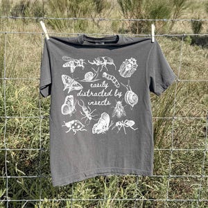 May include: A charcoal grey short-sleeve t-shirt featuring a white graphic print of various insects and snails surrounding the text "easily distracted by insects" in a cursive font, centred on the front of the garment.