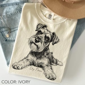 Könnte beinhalten: Ein elfenbeinfarbenes T-Shirt mit einer detaillierten schwarz-weißen Skizze eines liegenden Schnauzers. Das T-Shirt hat einen Rundhalsausschnitt und ein Comfort Colors Etikett. Unter der Hundeillustration steht der Text "Dog Name" in Schreibschrift.