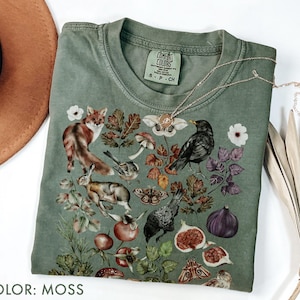Può includere: Una maglietta Comfort Colors color muschio con una grafica dettagliata a tema natura. Il design include una volpe, un coniglio, uccelli, funghi, fichi e falene. Una collana color oro è drappeggiata sulla maglietta.