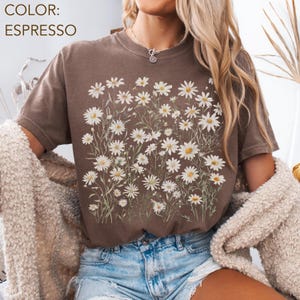 May include: Espresso-colored t-shirt featuring a floral design of white daisies with yellow centers and green stems. The text "COLOR: ESPRESSO" is visible in the upper left corner. The shirt has short sleeves and a crew neck.