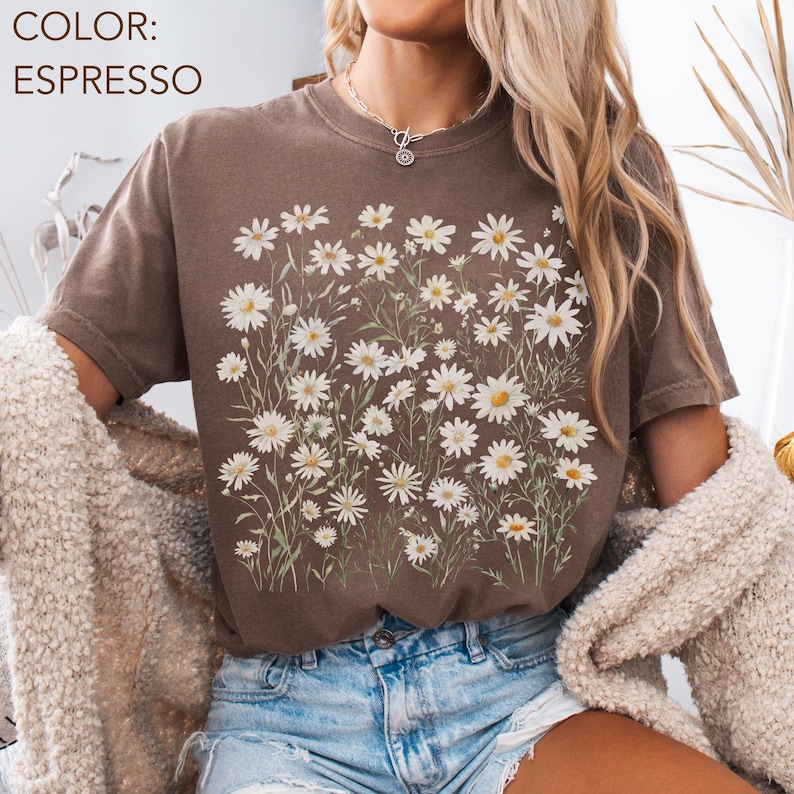 May include: Espresso-colored t-shirt featuring a floral design of white daisies with yellow centers and green stems. The text "COLOR: ESPRESSO" is visible in the upper left corner. The shirt has short sleeves and a crew neck.