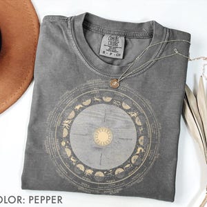 Pode incluir: Uma camiseta cinza Comfort Colors com um design dourado do zod&iacute;aco. O design apresenta um sol central e um arranjo circular de s&iacute;mbolos e texto do zod&iacute;aco. Um colar de prata com um pingente repousa sobre a camisa. A camisa est&aacute; dobrada, com um chap&eacute;u marrom e uma planta seca no fundo.