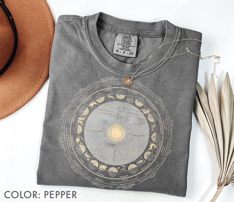 Pode incluir: Uma camiseta cinza Comfort Colors com um design dourado do zod&iacute;aco. O design apresenta um sol central e um arranjo circular de s&iacute;mbolos e texto do zod&iacute;aco. Um colar de prata com um pingente repousa sobre a camisa. A camisa est&aacute; dobrada, com um chap&eacute;u marrom e uma planta seca no fundo.