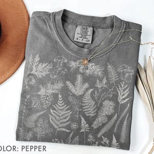 May include: A pepper-colored t-shirt with a botanical design featuring ferns, mushrooms, and other plants. The shirt has a crew neck and short sleeves. A silver necklace with a pendant is draped on the shirt. The text "COLOR: PEPPER" is at the bottom.
