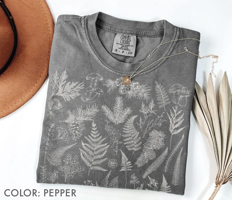 May include: A pepper-colored t-shirt with a botanical design featuring ferns, mushrooms, and other plants. The shirt has a crew neck and short sleeves. A silver necklace with a pendant is draped on the shirt. The text "COLOR: PEPPER" is at the bottom.
