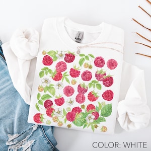 May include: White sweatshirt featuring a watercolor design of red raspberries, green leaves, and white blossoms. A silver necklace with a pendant rests on the shirt. The text "COLOR: WHITE" is at the bottom.