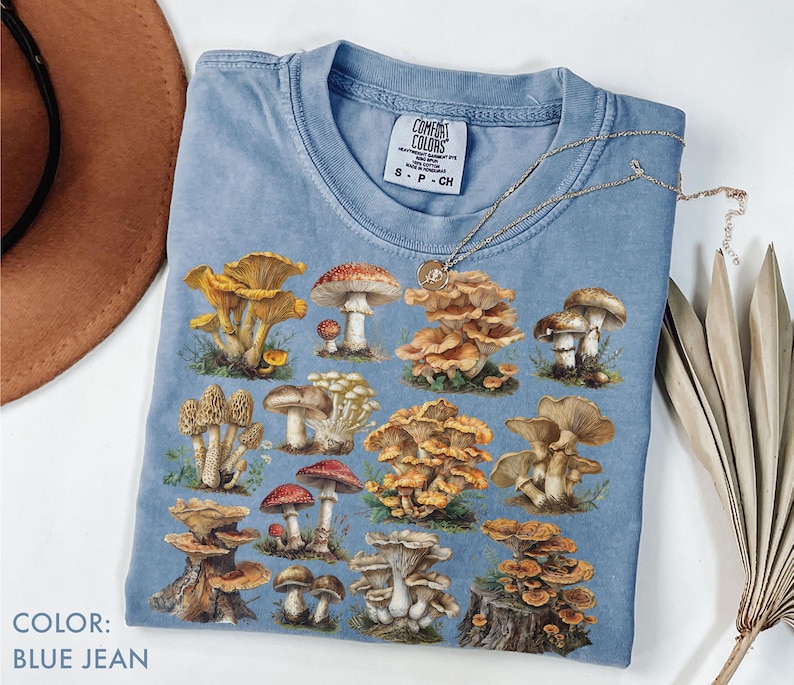 Pu&ograve; includere: T-shirt blu jeans con un'illustrazione dettagliata di vari funghi. La maglietta ha un girocollo e maniche corte. Una collana d'argento con un ciondolo &egrave; drappeggiata sulla maglietta. Il testo "COLOR: BLUE JEAN" &egrave; in basso a sinistra.