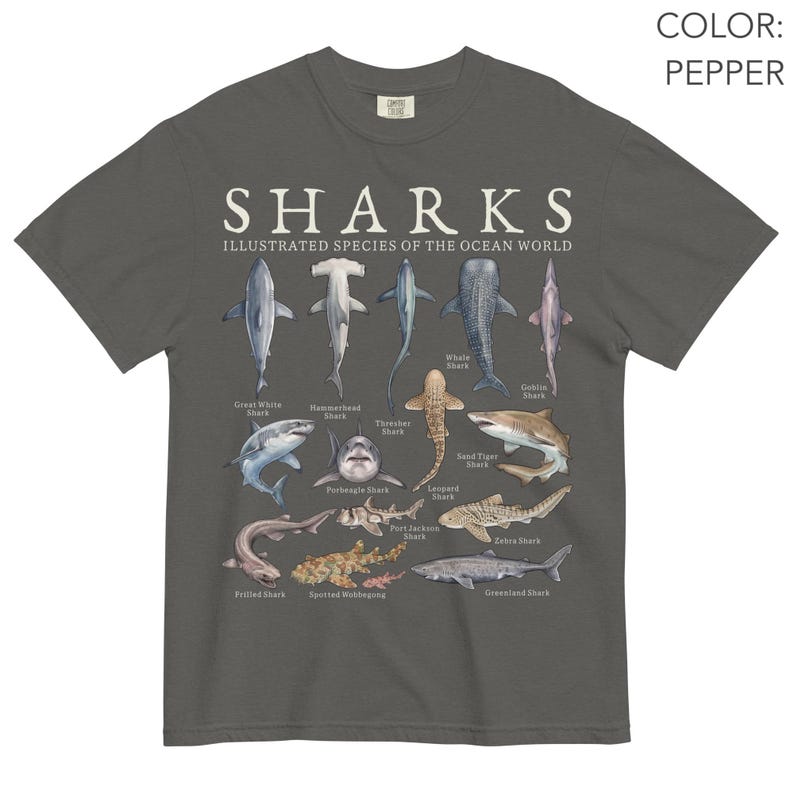 May include: A pepper-colored t-shirt featuring a graphic print titled SHARKS ILLUSTRATED SPECIES OF THE OCEAN WORLD. The design displays various shark species including the Great White, Hammerhead, Thresher, Whale, Goblin, Porbeagle, Sand Tiger, Frilled, Port Jackson, Leopard, Zebra, Spotted Wobbegong, and Greenland shark.