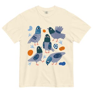 May include: Cream-colored t-shirt featuring a playful illustration of five pigeons in various poses. The birds are depicted in shades of gray and blue, with orange and blue accents. A soccer ball is also included in the design.