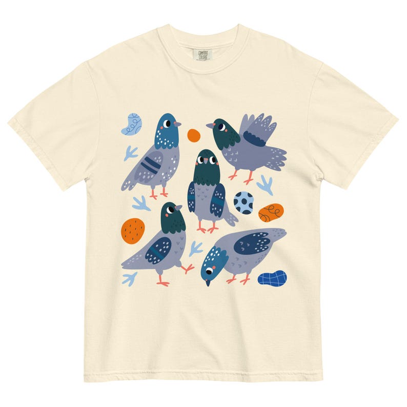 May include: Cream-colored t-shirt featuring a playful illustration of five pigeons in various poses. The birds are depicted in shades of gray and blue, with orange and blue accents. A soccer ball is also included in the design.