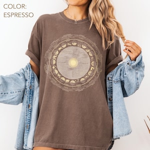 Pode incluir: T-shirt cor espresso com um design astrol&oacute;gico circular, com um sol no centro e s&iacute;mbolos do zod&iacute;aco. A camisa tem mangas curtas e um corte folgado. O texto "COLOR: ESPRESSO" est&aacute; no canto superior esquerdo.