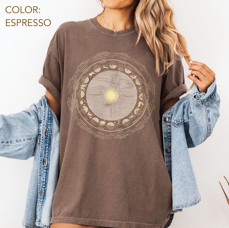 Pode incluir: T-shirt cor espresso com um design astrol&oacute;gico circular, com um sol no centro e s&iacute;mbolos do zod&iacute;aco. A camisa tem mangas curtas e um corte folgado. O texto "COLOR: ESPRESSO" est&aacute; no canto superior esquerdo.