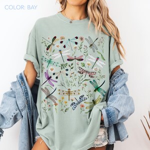 May include: A light green t-shirt with a colorful dragonfly and floral design. The shirt features various dragonflies in shades of purple, green, pink, and blue, surrounded by flowers and greenery. The text "COLOR: BAY" is at the top.
