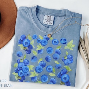 May include: Blue jean colored t-shirt with a blueberry design. The shirt features a crew neck and short sleeves. The design includes clusters of blue blueberries with green leaves. The shirt has a small tag on the upper left side. A gold necklace is draped across the shirt.