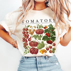 May include: Cream-colored t-shirt featuring a botanical illustration of various tomatoes in shades of red, green, and yellow. The word "TOMATOES" is printed above the illustration, with "SOLANUM LYCOPERSICUM - BOTANICAL STUDY" below.