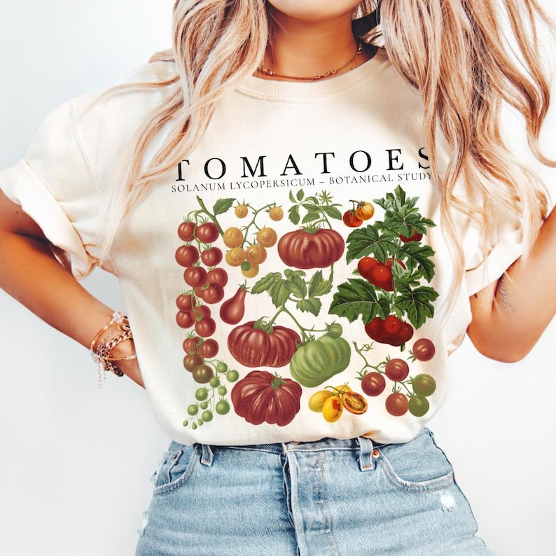 May include: Cream-colored t-shirt featuring a botanical illustration of various tomatoes in shades of red, green, and yellow. The word "TOMATOES" is printed above the illustration, with "SOLANUM LYCOPERSICUM - BOTANICAL STUDY" below.
