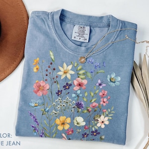 Puede incluir: Una camiseta azul vaquero con un dise&ntilde;o floral. La camiseta presenta un colorido ramo de flores de estilo acuarela en rosa, amarillo, morado y azul. Un collar de oro con un colgante descansa sobre la camiseta.