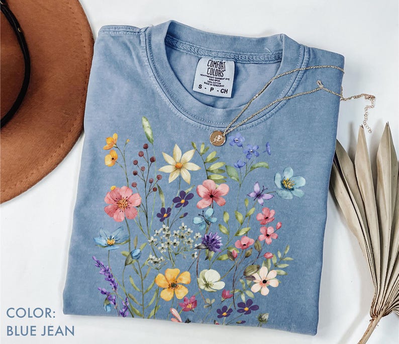 Puede incluir: Una camiseta azul vaquero con un dise&ntilde;o floral. La camiseta presenta un colorido ramo de flores de estilo acuarela en rosa, amarillo, morado y azul. Un collar de oro con un colgante descansa sobre la camiseta.