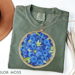 May include: A moss green t-shirt featuring a watercolor illustration of a bowl overflowing with blueberries. The shirt has a crew neck and short sleeves. A gold necklace with a small pendant rests on the shirt. The text "COLOR: MOSS" is at the bottom.