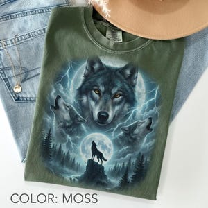 K&ouml;nnte beinhalten: Moosgr&uuml;nes T-Shirt mit detaillierter Grafik von W&ouml;lfen, die den Mond anheulen, mit Blitzen und Waldlandschaft. Das Shirt hat einen Rundhalsausschnitt und kurze &Auml;rmel. Der Text "COLOR: MOSS" steht unten.