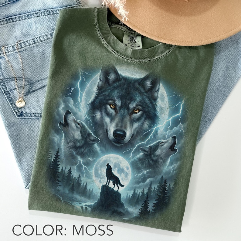K&ouml;nnte beinhalten: Moosgr&uuml;nes T-Shirt mit detaillierter Grafik von W&ouml;lfen, die den Mond anheulen, mit Blitzen und Waldlandschaft. Das Shirt hat einen Rundhalsausschnitt und kurze &Auml;rmel. Der Text "COLOR: MOSS" steht unten.