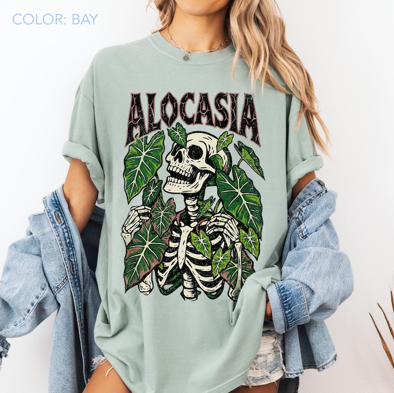 K&ouml;nnte beinhalten: Hellgr&uuml;nes T-Shirt mit dem Wort "ALOCASIA" in dunkelroter, gotischer Schrift. Die Grafik zeigt ein Skelett, das von gr&uuml;nen Bl&auml;ttern umgeben ist. Das T-Shirt hat eine lockere Passform.