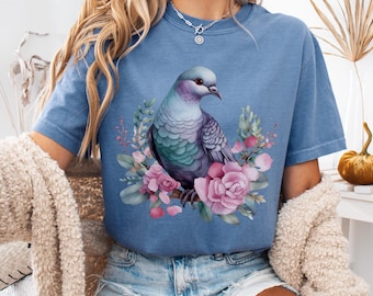 Könnte beinhalten: Blaues Jeans-T-Shirt mit einer Aquarellillustration einer Taube, umgeben von rosa Rosen und Grün. Der Text "COLOR: BLUE JEAN" ist sichtbar. Das Shirt hat kurze Ärmel und einen Rundhalsausschnitt.