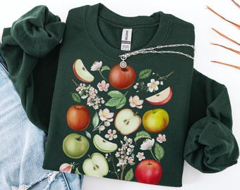May include: Forest green sweatshirt featuring a colorful apple design with whole apples, apple slices, and blossoms. A silver necklace with a small charm is draped across the sweatshirt. The text "COLOR: FOREST GREEN" is at the bottom.