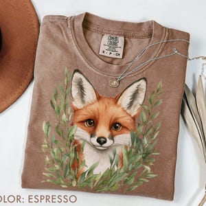 Peut inclure: T-shirt couleur expresso avec une illustration détaillée du visage d'un renard entouré de feuilles vertes. Le t-shirt a un col rond et des manches courtes. Un collier argenté avec un petit pendentif est drapé sur le t-shirt.