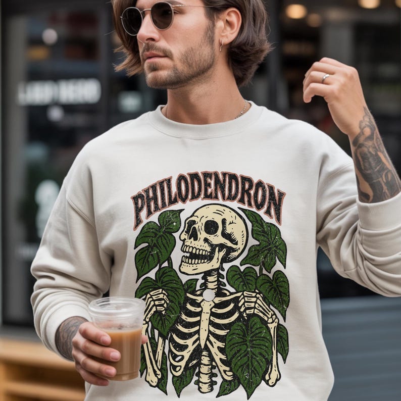 May include: Cream-colored sweatshirt with a graphic featuring a skeleton surrounded by green leaves, with the word "PHILODENDRON" arched above. The design has a vintage, slightly distressed look.