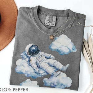 Peut inclure: T-shirt couleur poivre avec une illustration &agrave; l'aquarelle d'un astronaute se relaxant sur des nuages. L'astronaute porte une combinaison spatiale et un casque blancs. Le t-shirt a un col rond et des manches courtes. Le texte "COLOR: PEPPER" est en bas.