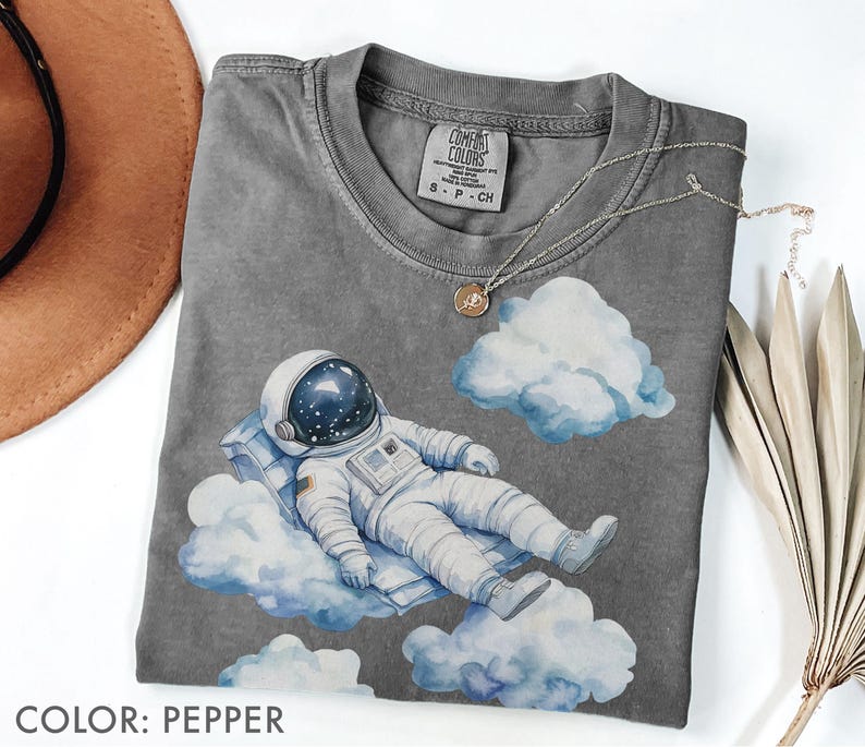 Peut inclure: T-shirt couleur poivre avec une illustration &agrave; l'aquarelle d'un astronaute se relaxant sur des nuages. L'astronaute porte une combinaison spatiale et un casque blancs. Le t-shirt a un col rond et des manches courtes. Le texte "COLOR: PEPPER" est en bas.