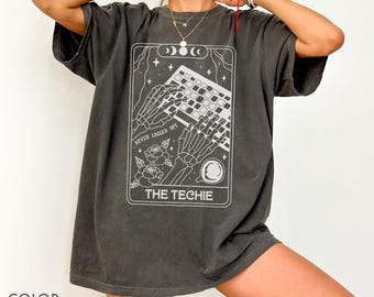 May include: Pepper-colored t-shirt featuring a white graphic of skeleton hands typing on a keyboard, with the text "NEVER LOGGED OFF" and "THE TECHIE". The design includes moons, stars, and roses. The shirt is a relaxed fit.