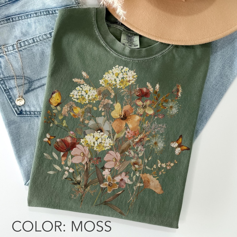 May include: A moss-colored t-shirt featuring a floral design with butterflies. The graphic includes a variety of flowers in shades of white, yellow, pink, and orange. The text "COLOR: MOSS" is at the bottom of the image.
