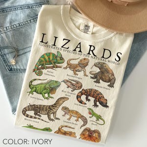 May include: An ivory t-shirt featuring the text LIZARDS ILLUSTRATED SPECIES OF THE NATURAL WORLD and detailed illustrations of various lizard species including a veiled chameleon, bearded dragon, komodo dragon, green iguana, and leopard gecko.