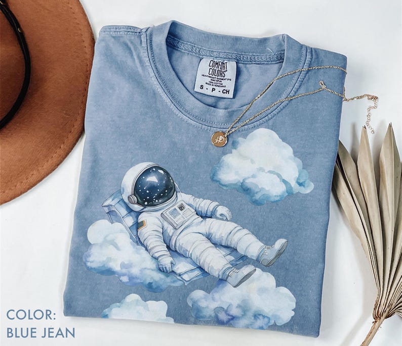 Peut inclure: Un t-shirt bleu jean avec un motif d'astronaute flottant sur des nuages. L'astronaute porte une combinaison spatiale et un casque blancs. Le t-shirt a une &eacute;tiquette Comfort Colors. Un collier en or avec un pendentif est sur le t-shirt.