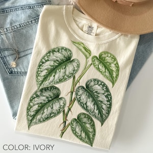 May include: Ivory-colored Comfort Colors t-shirt featuring a botanical illustration of a plant with large, green and white patterned leaves. The shirt is paired with light blue jeans and a gold necklace. The text "COLOR: IVORY" is at the bottom.
