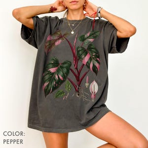 Peut inclure: T-shirt gris poivre avec une illustration botanique d'une plante aux feuilles et tiges roses et vertes. L'illustration comprend une fleur et des racines.