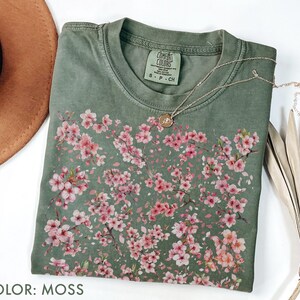 May include: A moss green t-shirt with a floral design of pink blossoms. The shirt has a crew neck and short sleeves. A gold necklace with a small pendant is draped across the shirt. The text "COLOR: MOSS" is at the bottom of the image.