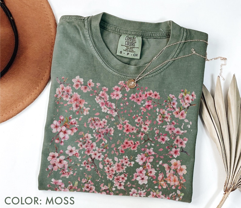 May include: A moss green t-shirt with a floral design of pink blossoms. The shirt has a crew neck and short sleeves. A gold necklace with a small pendant is draped across the shirt. The text "COLOR: MOSS" is at the bottom of the image.
