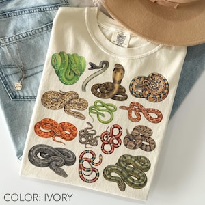 Könnte beinhalten: Ein elfenbeinfarbenes T-Shirt mit einem Grafikdruck verschiedener Schlangen in mehreren Farben, darunter Grün, Braun, Orange, Rot und Schwarz. Das T-Shirt hat einen Rundhalsausschnitt und ein Comfort Colors Markenetikett im Kragenbereich.
