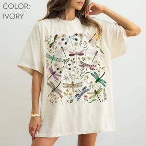 May include: Ivory-colored oversized t-shirt featuring a colorful dragonfly and floral print. The design includes various dragonflies in shades of green, purple, pink, and blue, along with small flowers and greenery. The shirt has a relaxed fit.