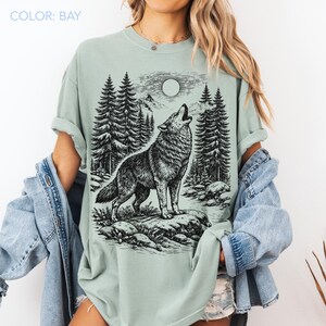 Puede incluir: Una camiseta color bah&iacute;a con un gr&aacute;fico en tinta negra de un lobo aullando a la luna llena en un entorno forestal. El dise&ntilde;o incluye pinos altos, terreno rocoso y picos de monta&ntilde;as. La camiseta tiene cuello redondo y mangas cortas.