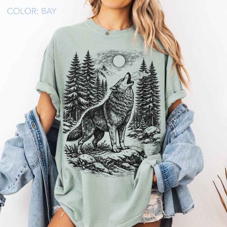 Puede incluir: Una camiseta color bah&iacute;a con un gr&aacute;fico en tinta negra de un lobo aullando a la luna llena en un entorno forestal. El dise&ntilde;o incluye pinos altos, terreno rocoso y picos de monta&ntilde;as. La camiseta tiene cuello redondo y mangas cortas.