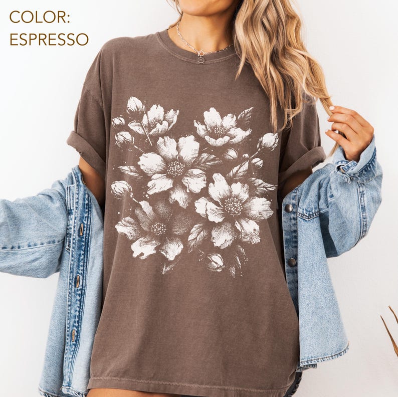 以下が含まれることがあります： 大きな白い花の模様が特徴の、エスプレッソ色のTシャツ。クルーネックと半袖です。「COLOR: ESPRESSO」の文字が左上にあります。明るいブルーのデニムジャケットと合わせています。