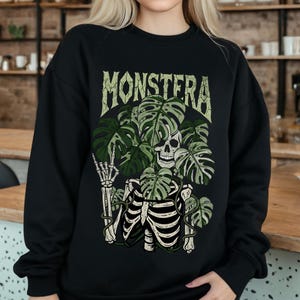 Può includere: Felpa nera con un design grafico raffigurante uno scheletro, foglie di monstera e la parola "MONSTERA" in un font invecchiato. Il design incorpora colori verdi e bianco sporco.